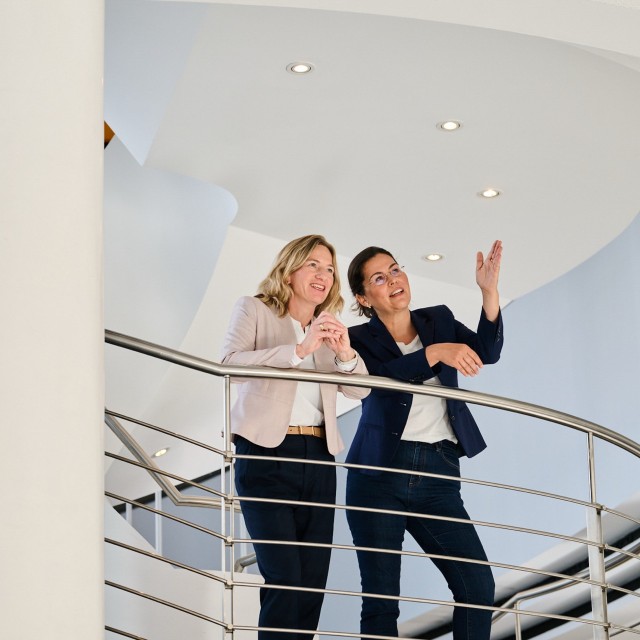 Zwei Frauen auf einer Wendeltreppe an ein Metallgeländer gelehnt, die sich unterhalten.