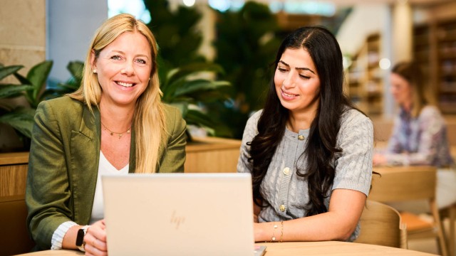 Zwei Frauen sitzen an einem Holztisch vor einem geöffneten Laptop.