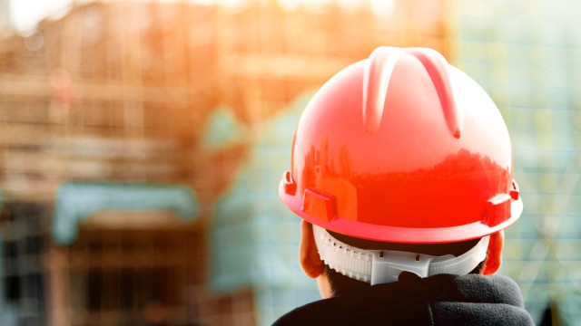 Der Hinterkopf eines Mannes der einen Baustellenhelm trägt. Im Hintergrund eine Baustelle, auf die er blickt.