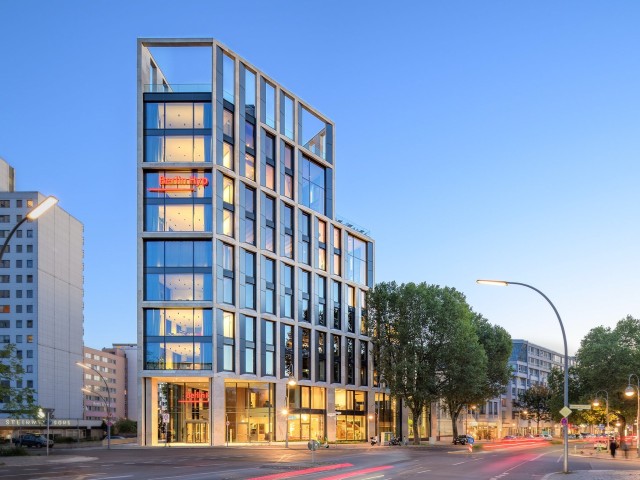 Die Außenfassade der von innen beleuchteten Unternehmenszentrale der Berlin Hyp bei Dämmerung, mit blauem Himmel.