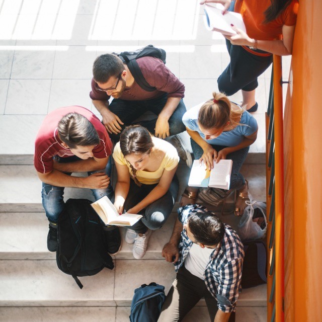 Vier Personen sitzen auf einer Treppe, zwei halten offene Bücher, drei lesen gemeinsam aus einem der Bücher.
