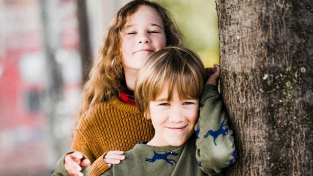 Zwei Kinder stehen hintereinander an einem Baum. Das hintere Kind ist etwa größer und schauen beide in die Kamera.